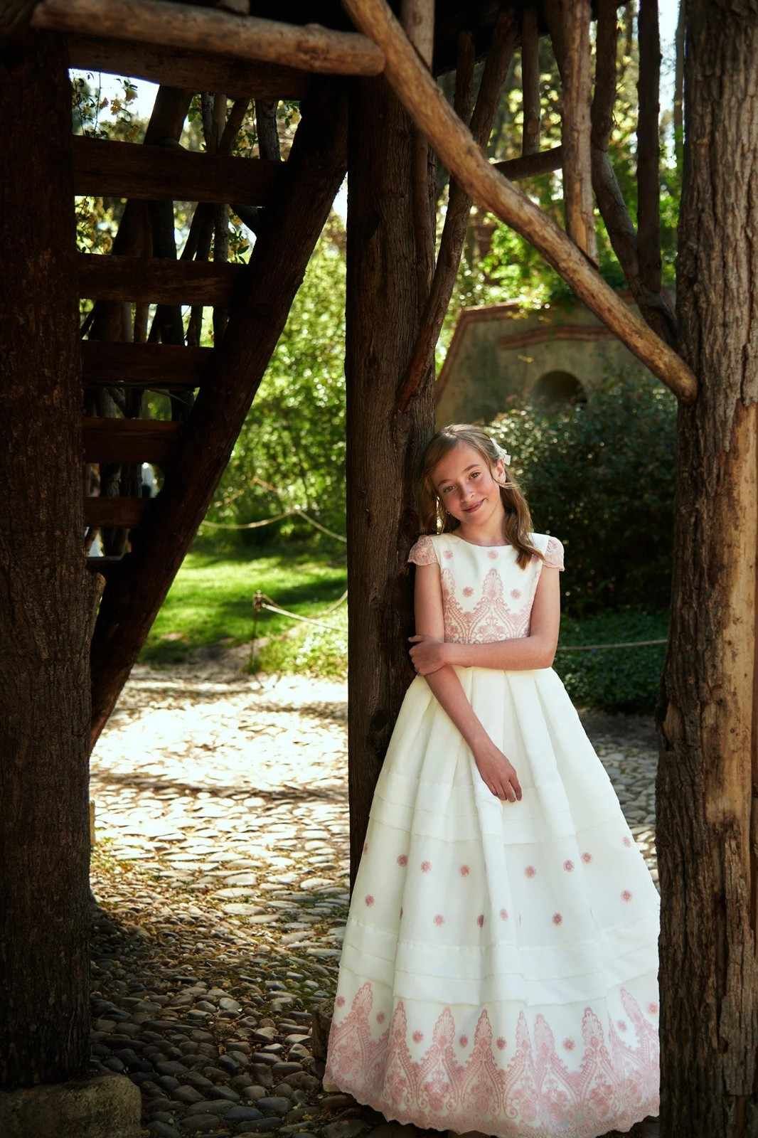 Mujer con vestido blanco de pie bajo una estructura de madera en un jardín soleado.