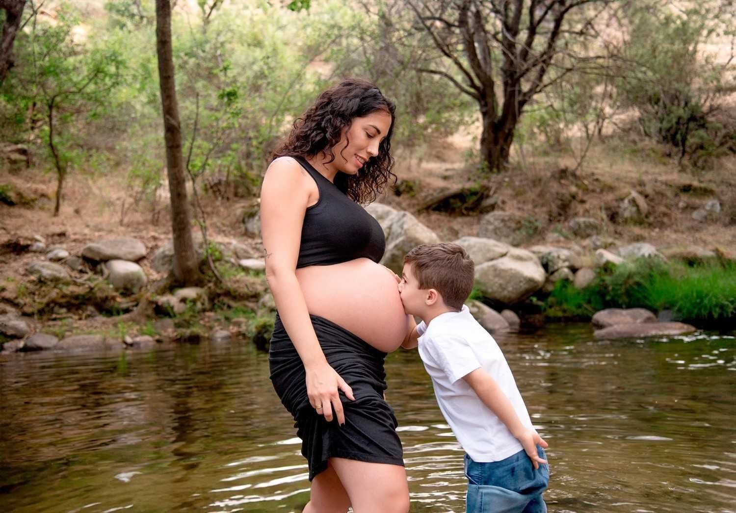 Una mujer embarazada vestida de negro está de pie junto a un arroyo mientras un niño le besa la barriga.