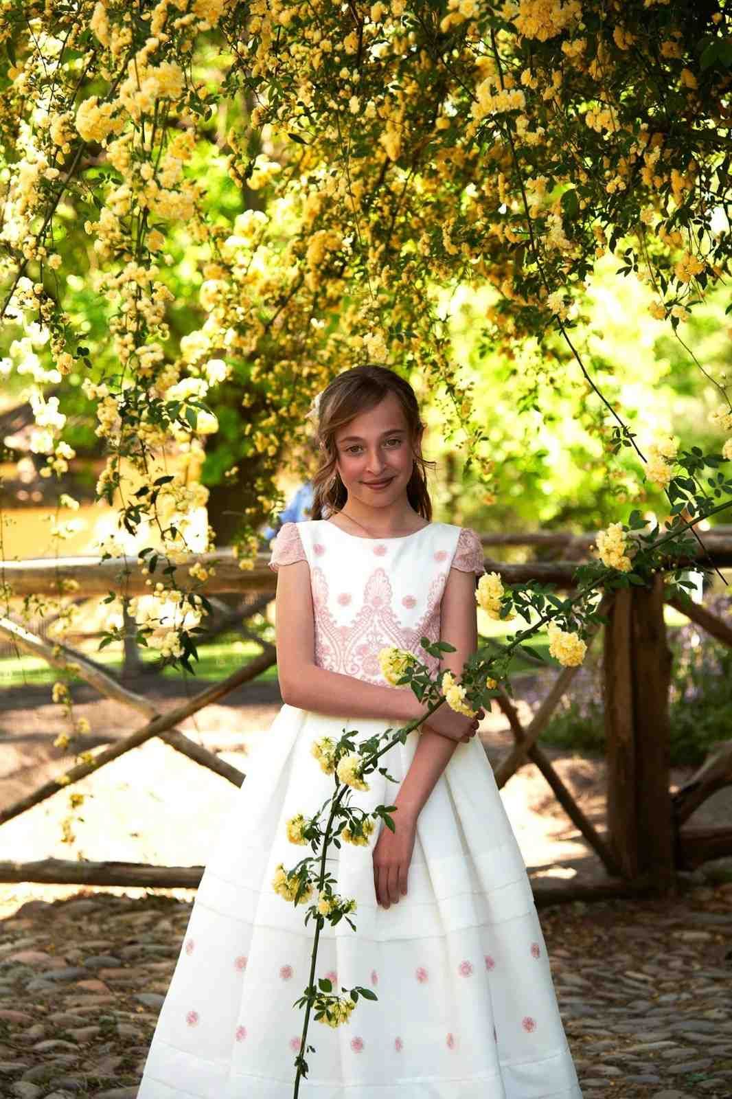 Una niña con un vestido blanco está de pie bajo unas ramas floridas al aire libre, sosteniendo una enredadera y sonriendo.