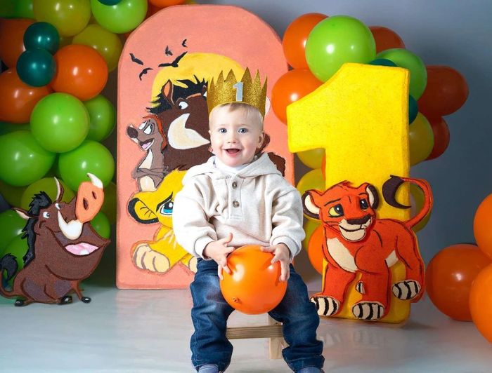 Un niño pequeño sostiene una pelota naranja en un colorido escenario de fiesta decorado con globos y figuras de dibujos animados.