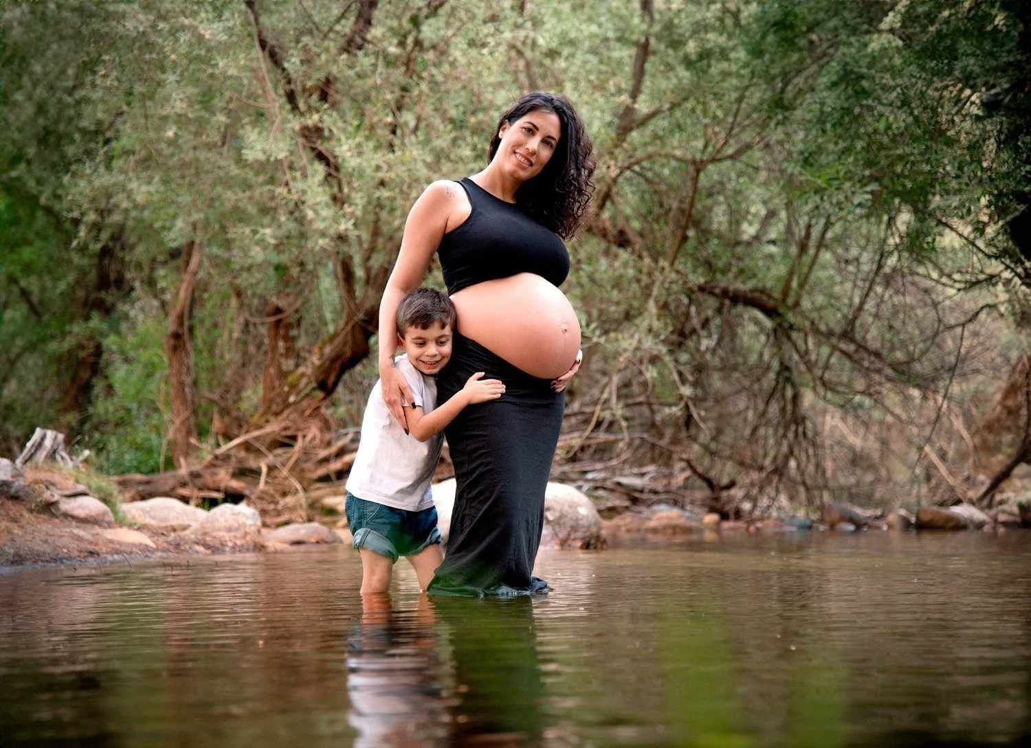 Una mujer embarazada se encuentra de pie en aguas poco profundas con un niño, sujetándose el vientre en un arroyo boscoso.