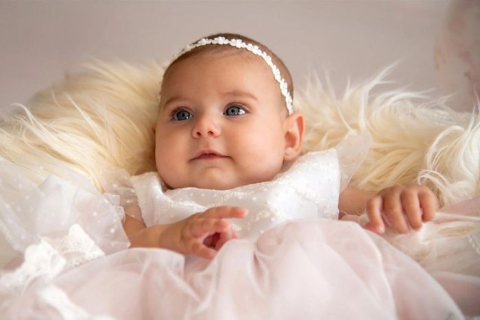 Bebé con un vestido blanco, tumbada sobre una suave manta color crema, con una delicada diadema.