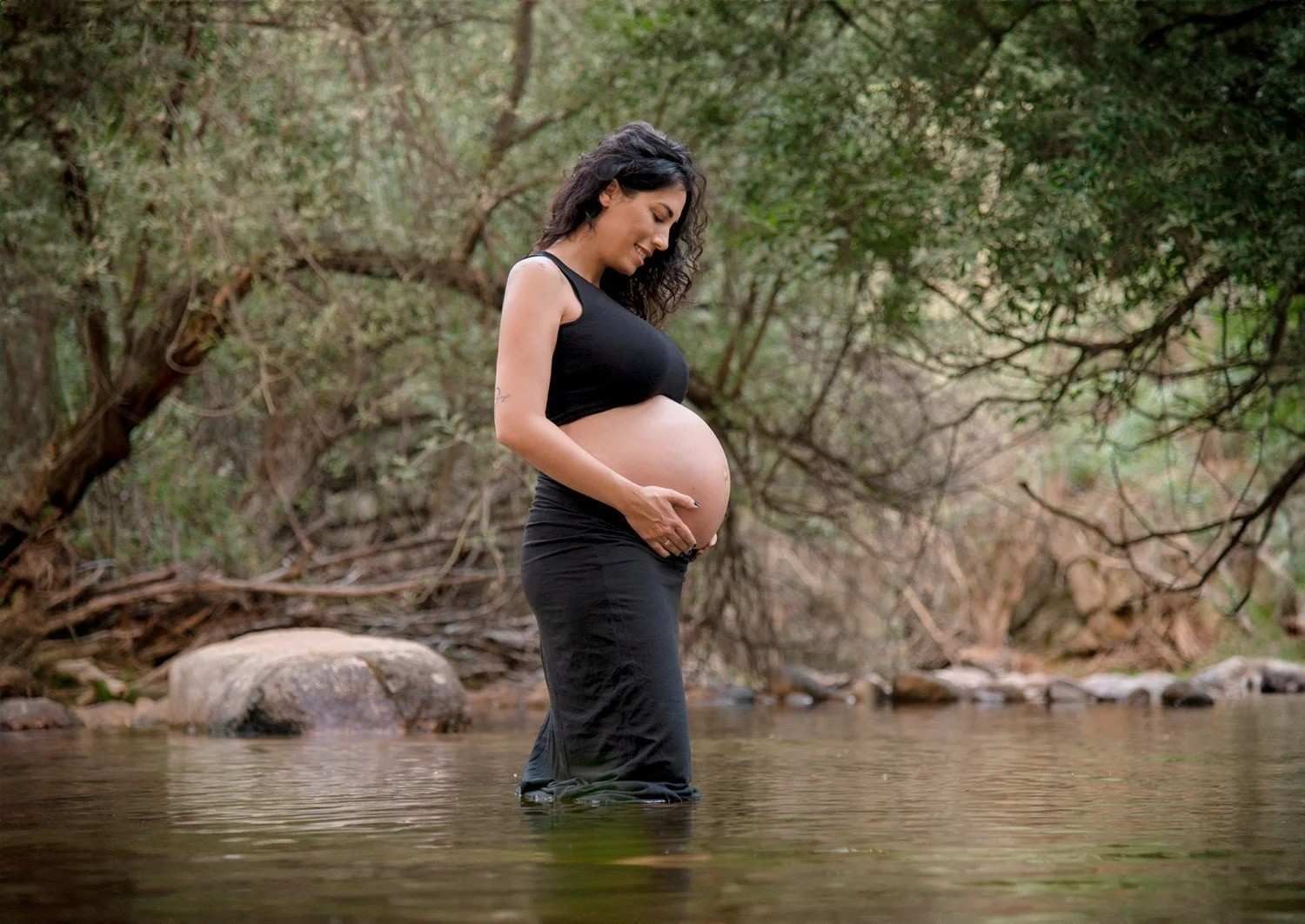 Mujer embarazada de pie en aguas poco profundas junto a árboles y rocas, sujetándose el vientre.