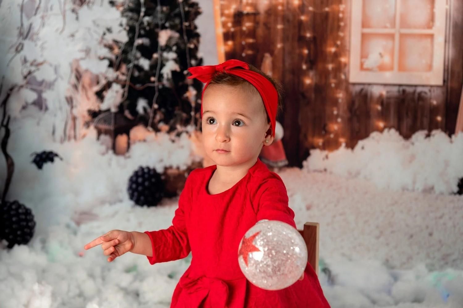 Niña pequeña con vestido rojo y diadema sosteniendo un adorno transparente en un entorno navideño nevado.