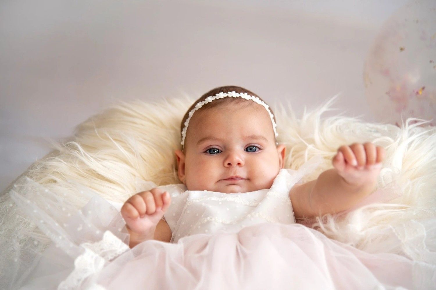 Bebé con un vestido blanco, tumbada sobre una manta suave, con una diadema de perlas y mirando a la cámara.
