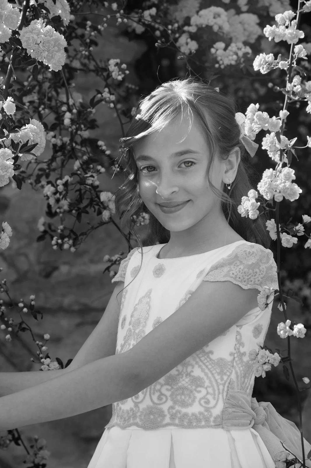 Niña sonriente con un vestido blanco de pie frente a un jardín florido.