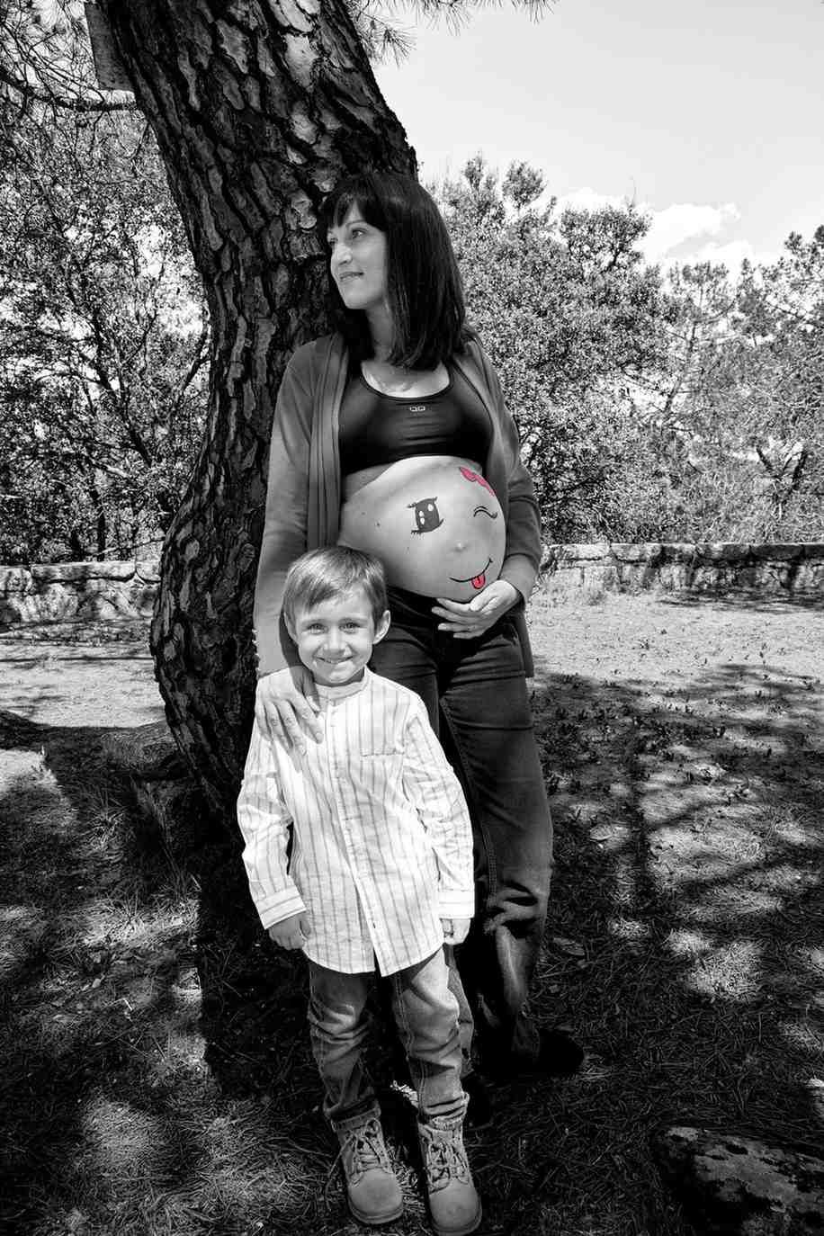 Una mujer embarazada y un niño pequeño de pie junto a un árbol en un parque, ambos mirando a la cámara.