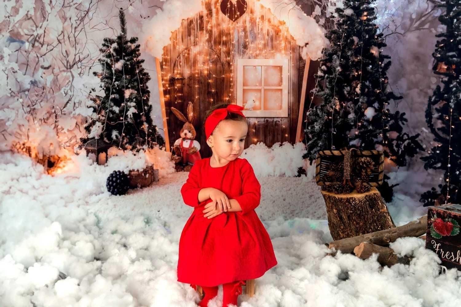 Niña con vestido rojo y gorro de Papá Noel de pie en una escena navideña nevada con árboles iluminados y una acogedora cabaña de fondo.
