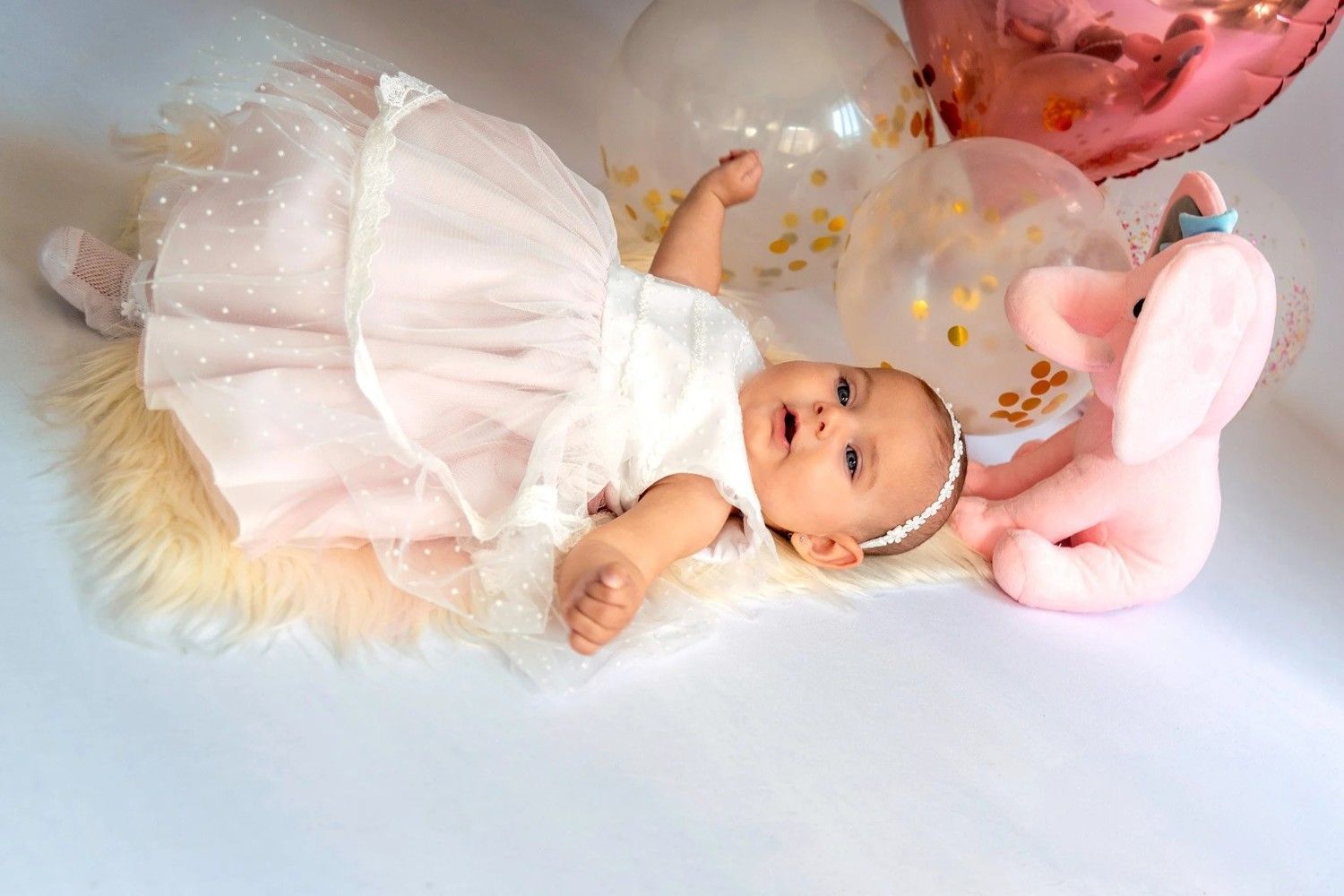 Bebé con un vestido blanco acostado sobre una manta suave con globos rosas y transparentes cerca