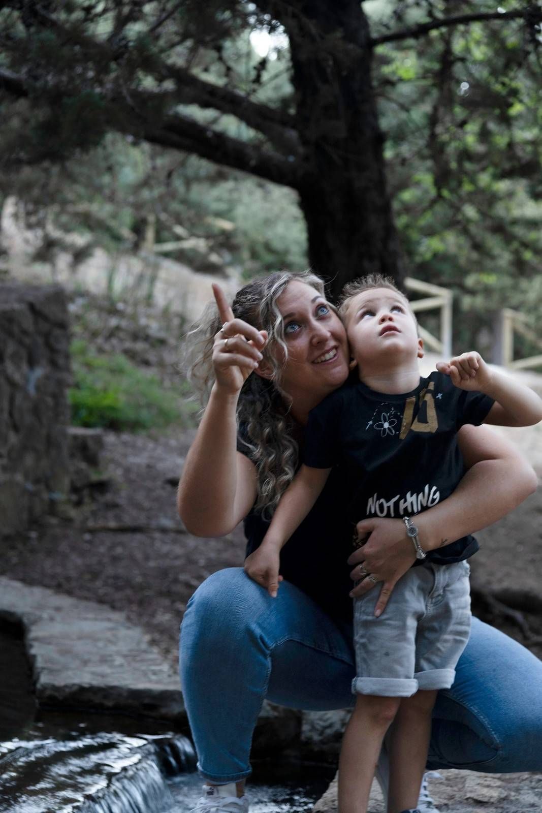 Mujer arrodillada junto a un arroyo, abrazando a un niño y señalando hacia arriba en un entorno boscoso al aire libre.