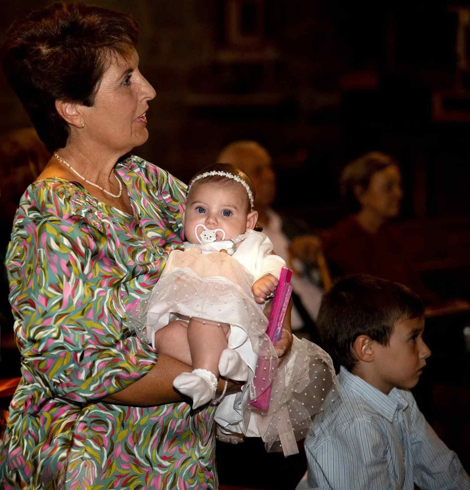 Una mujer vestida de blanco sostiene a un bebé mientras un niño permanece de pie cerca en un auditorio con poca luz.