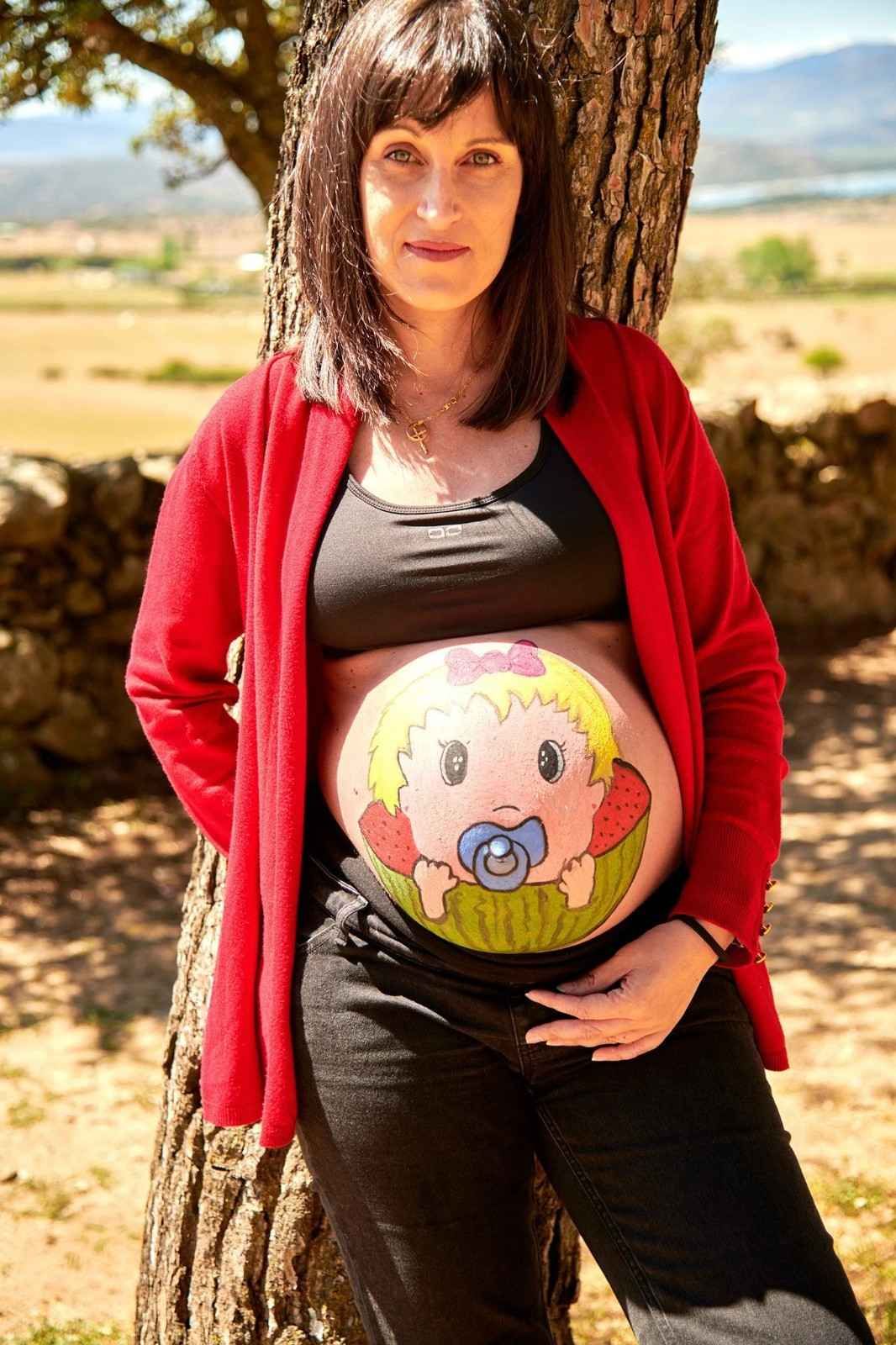 Mujer embarazada con cárdigan rojo de pie junto a un árbol en un campo soleado, sosteniendo su vientre pintado con una cara de dibujos animados.