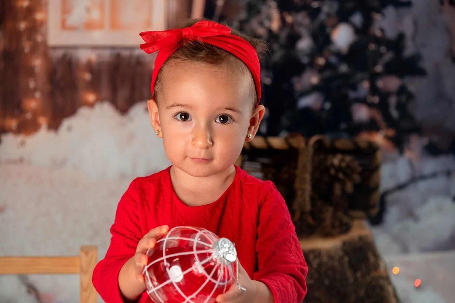 Niña con un atuendo rojo y una diadema, sosteniendo un adorno rojo, de pie en un entorno navideño con iluminación tenue.