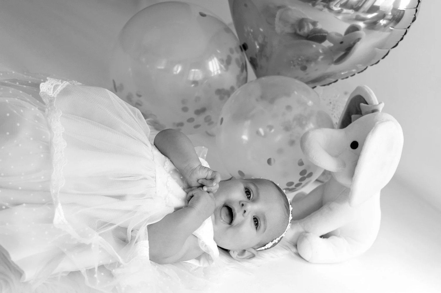 Un bebé con un vestido blanco yace entre globos junto a un elefante de peluche blanco.