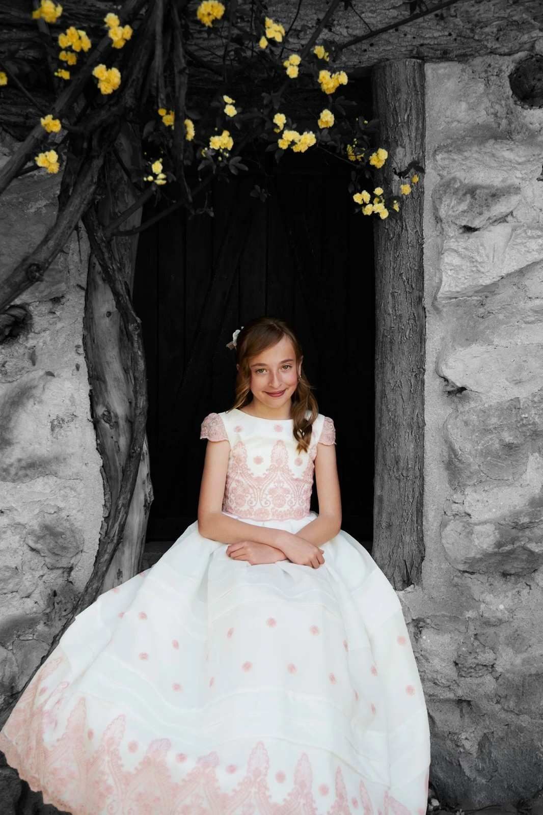 Niña con vestido blanco sentada en un portal de piedra bajo flores amarillas