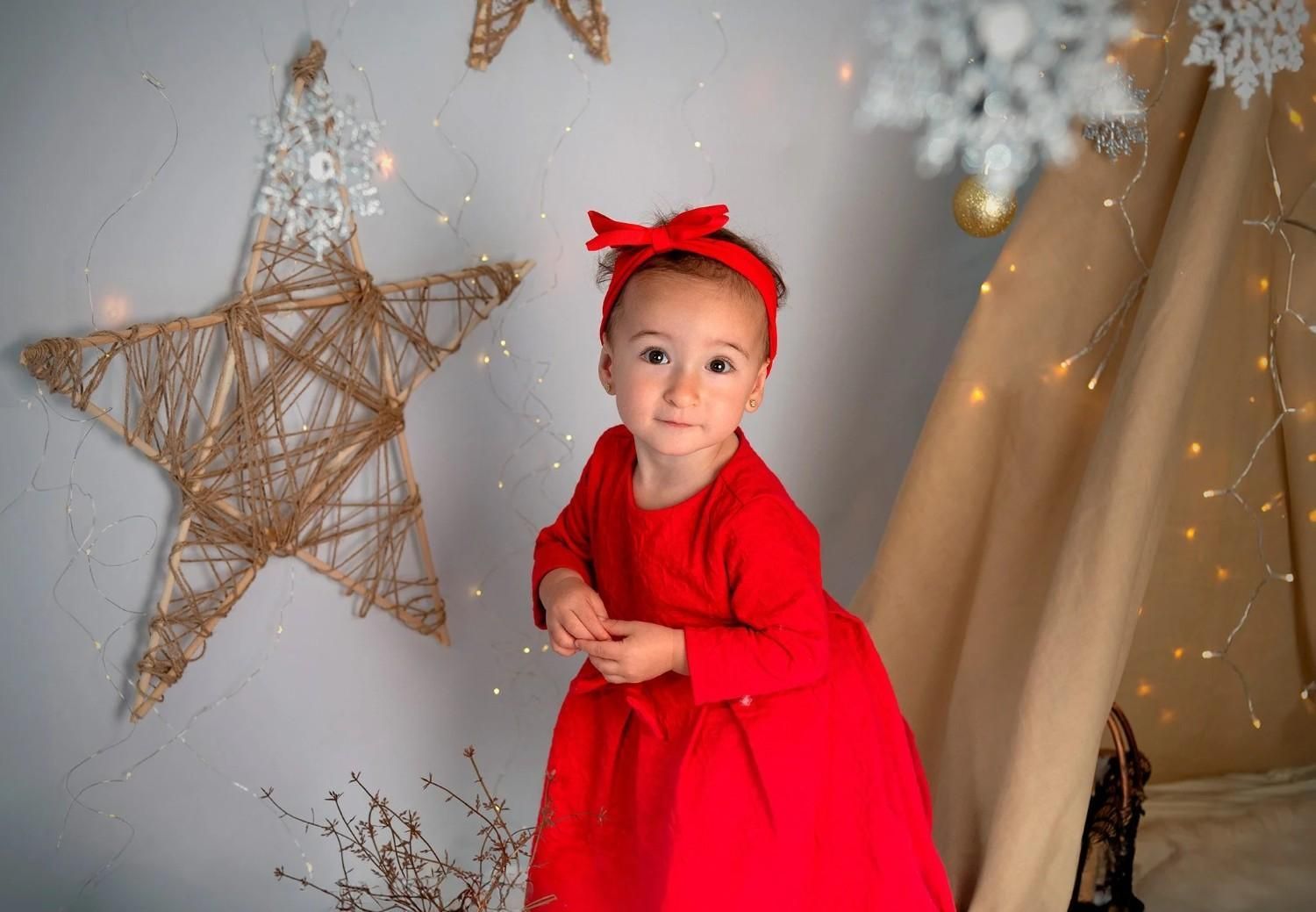 Niña con vestido rojo y diadema posando junto a una gran estrella decorativa y luces festivas.