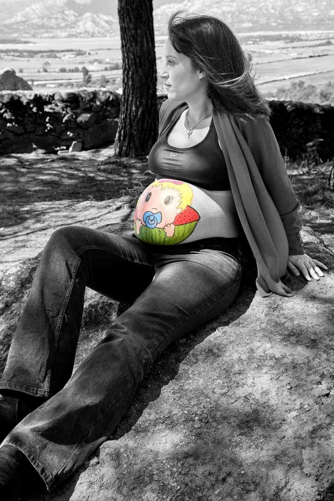 Una mujer embarazada sentada al aire libre junto a un árbol, descansando con el vientre pintado de colores.