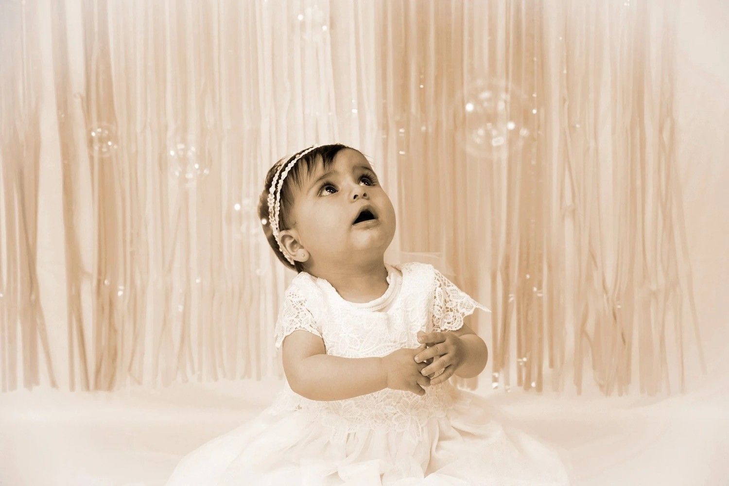 Niña pequeña con un vestido blanco mirando hacia arriba contra un fondo de cortina de color melocotón suave.