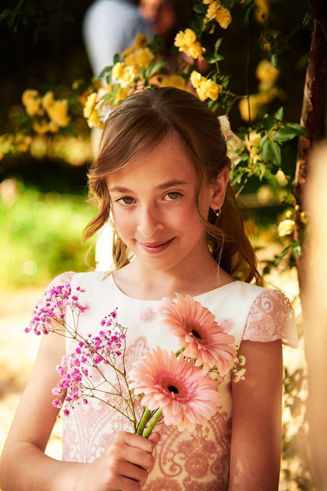 Una niña sostiene flores rosas en un jardín soleado, viste un vestido blanco y sonríe dulcemente.