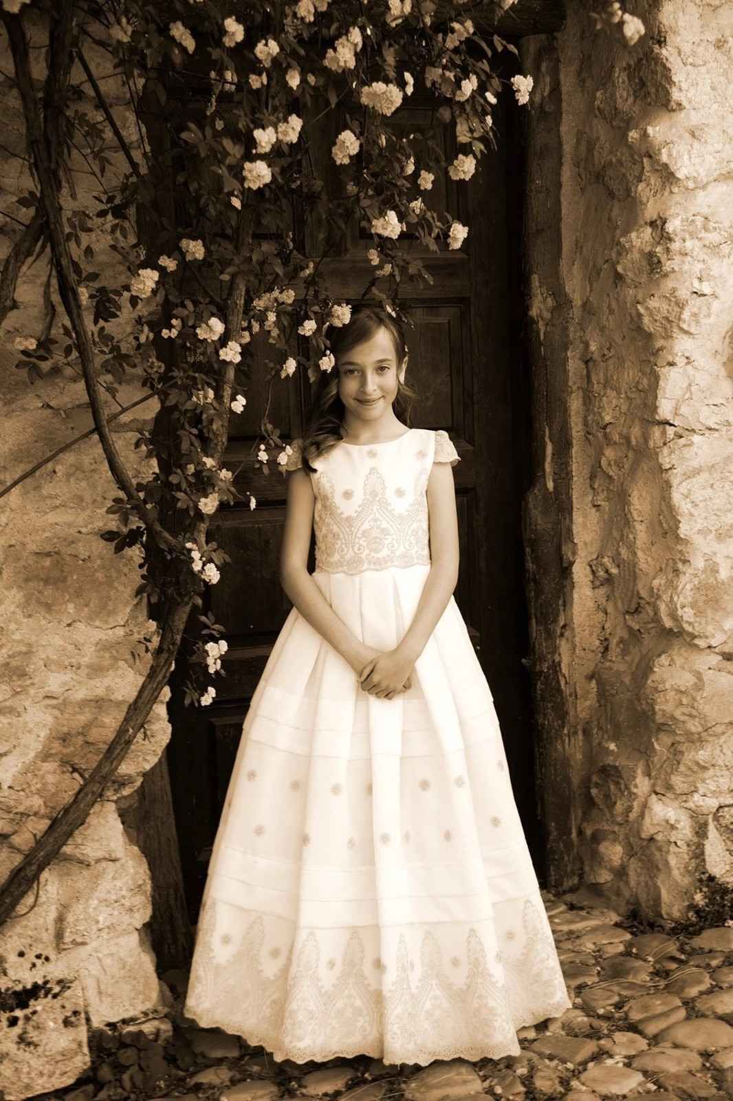 Una niña con un vestido blanco está de pie junto a una puerta de piedra cubierta de flores.