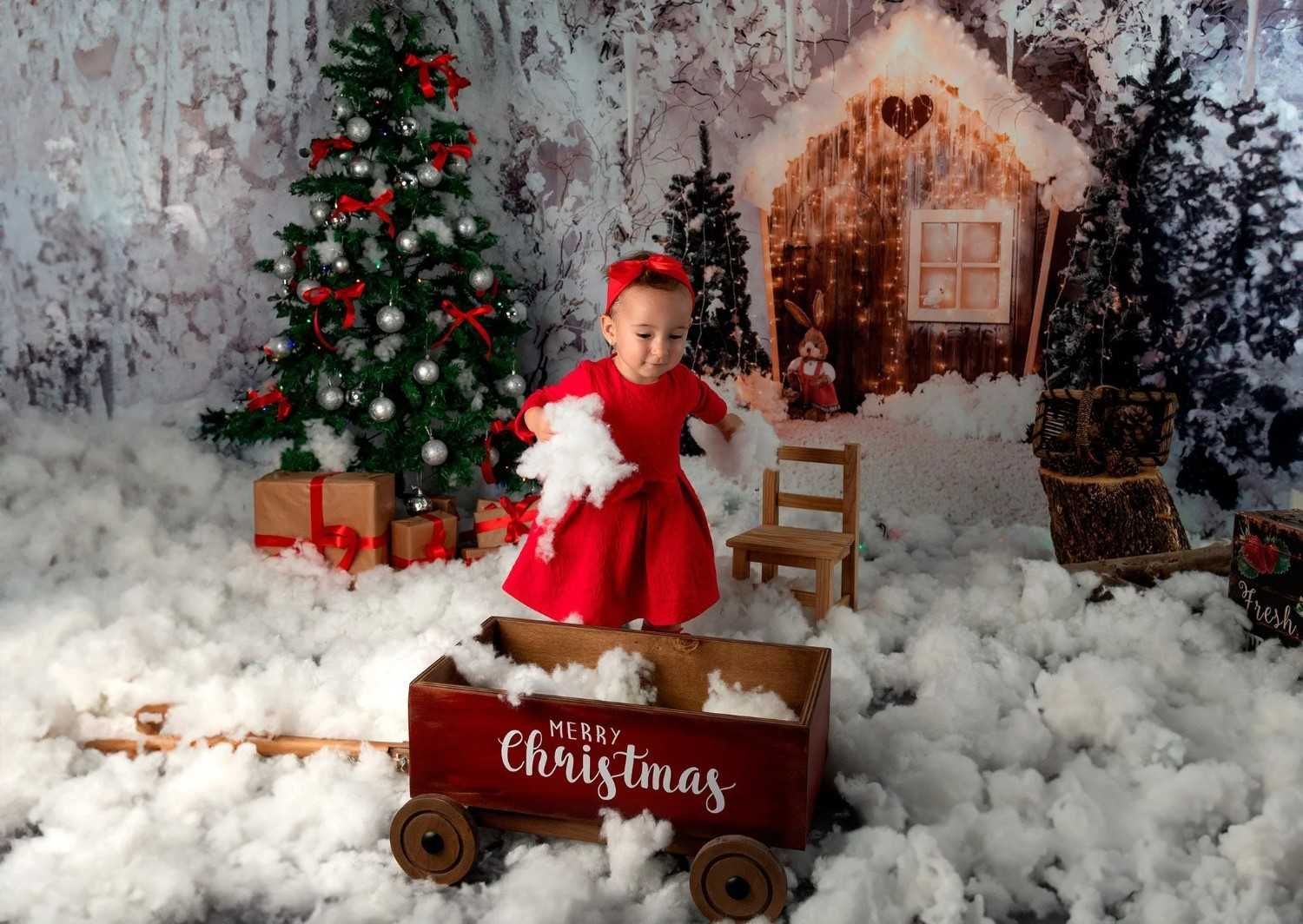 Niña con vestido rojo en una escena navideña nevada junto a un carrito con la etiqueta 
