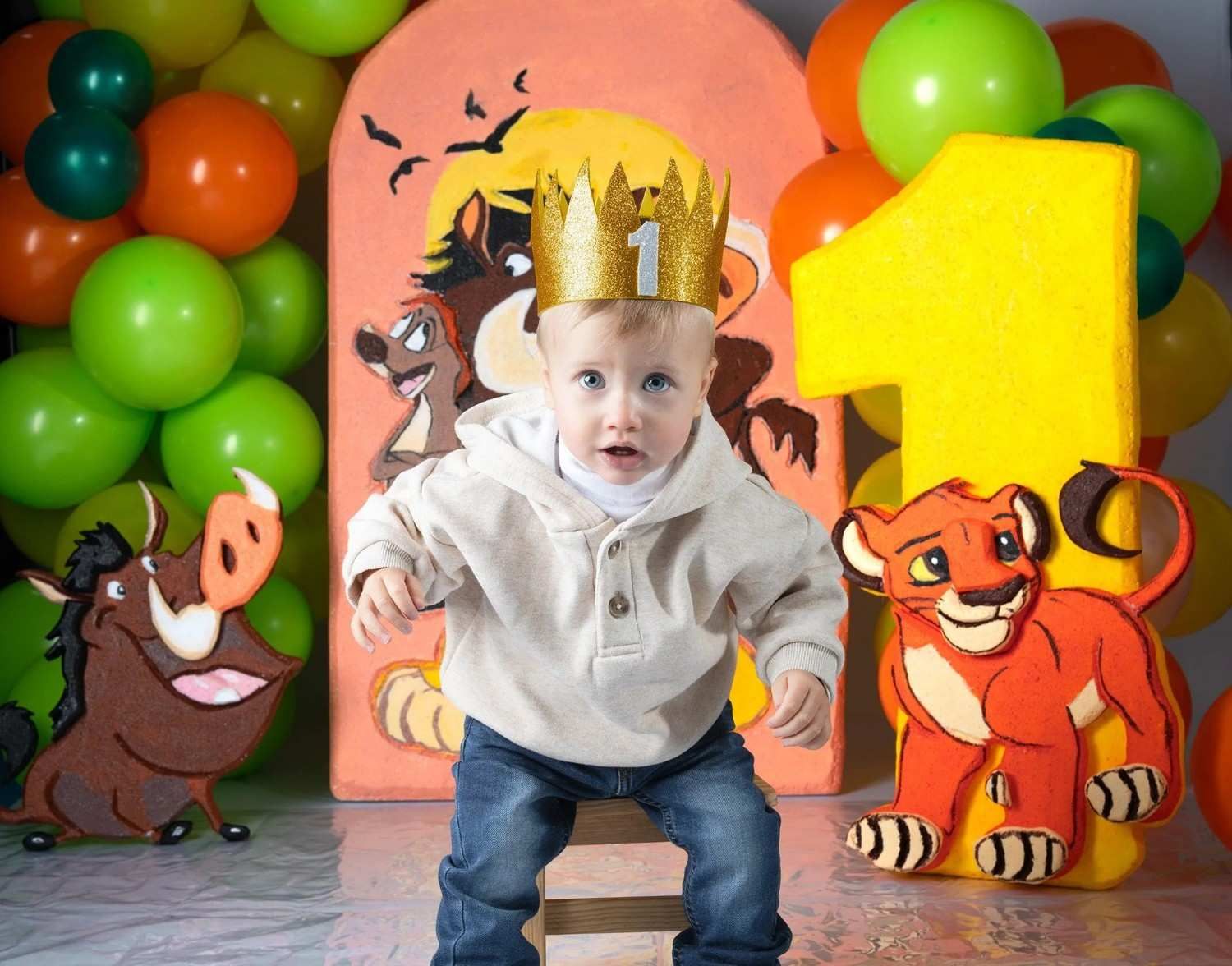 Un niño pequeño con una corona dorada da un paso al frente frente a un colorido telón de fondo de una fiesta con temática del Rey León.