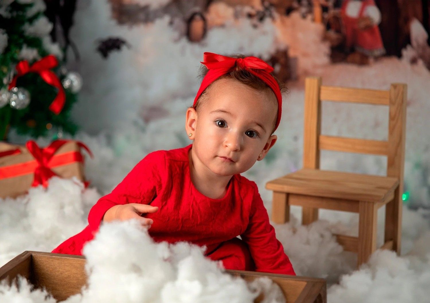 Bebé con conjunto rojo y diadema sentado en una decoración blanca y esponjosa junto a una pequeña silla de madera y adornos navideños.