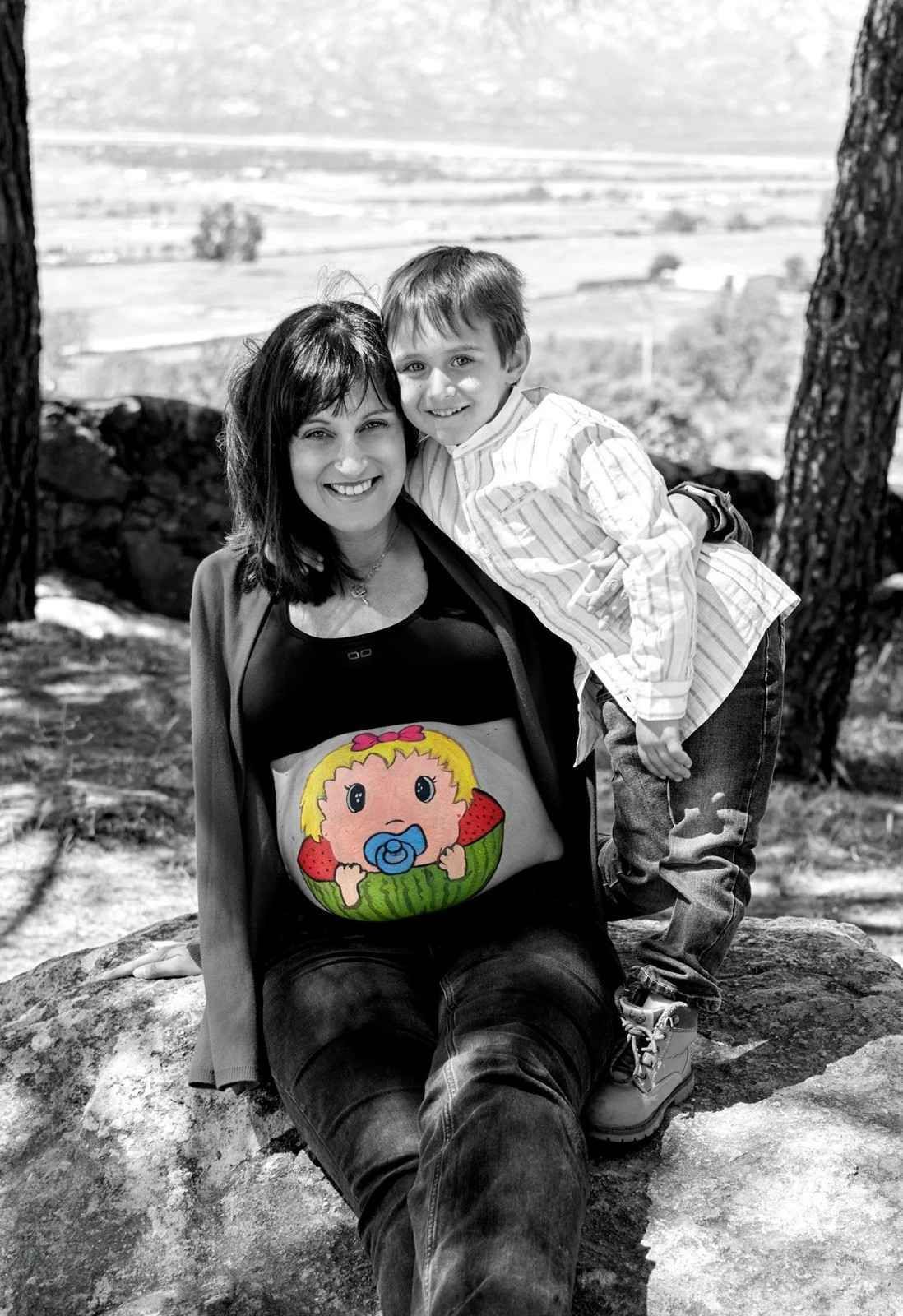 Una mujer sentada al aire libre con un niño apoyado en ella, ambos sonriendo cerca de un mirador panorámico.