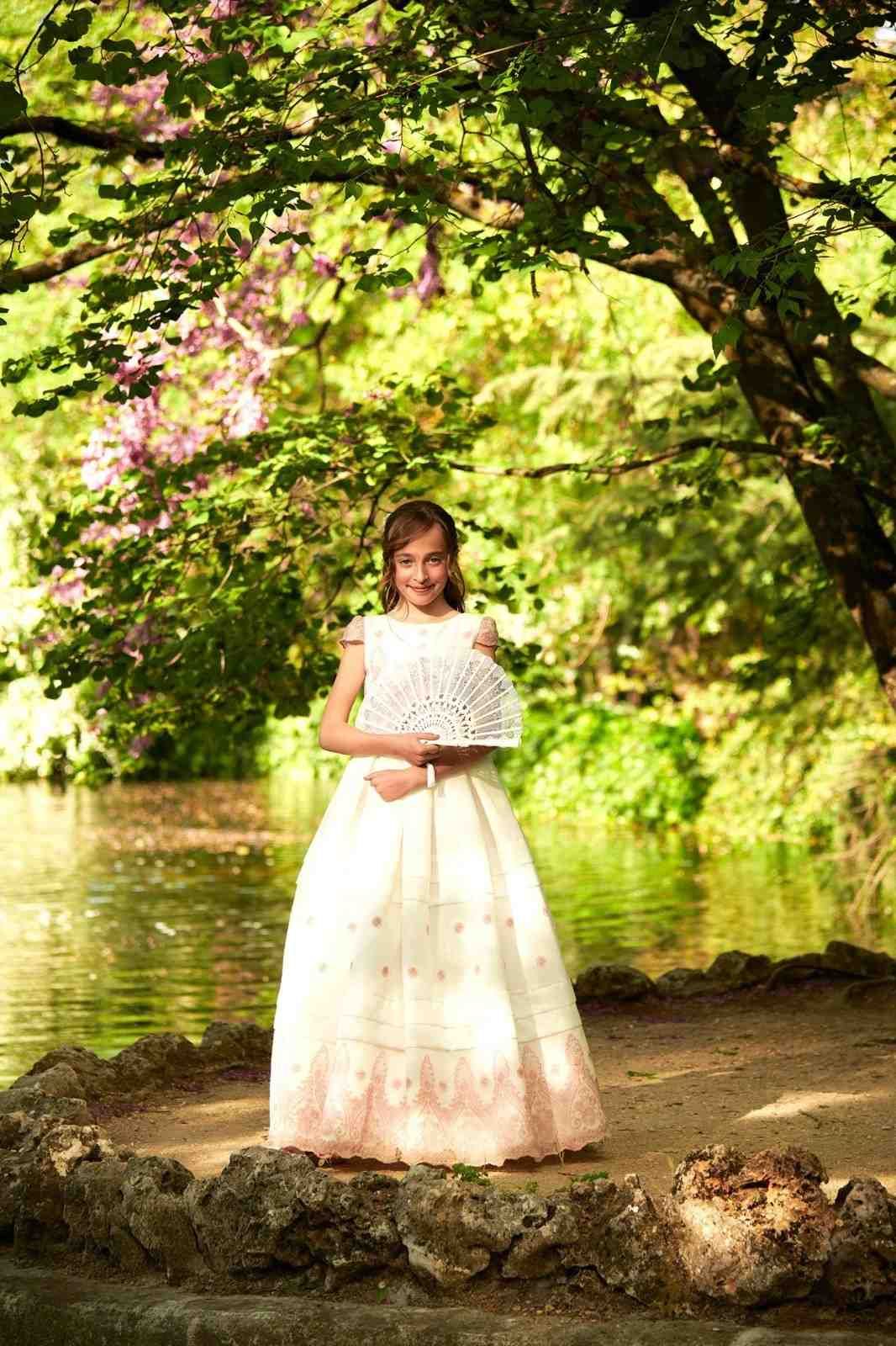 Mujer con vestido blanco de pie junto a un río bajo árboles en flor.