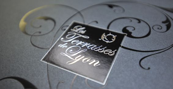 Détail d'un support de communication sur fond gris, avec lettrage argenté et volutes.