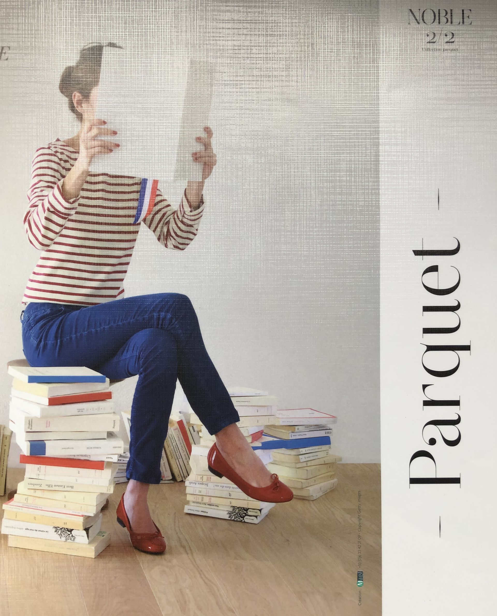 Femme lisant, assise sur des livres, vêtue d'un jean, de chaussures rouges et d'une chemise à rayures. Sol en parquet.