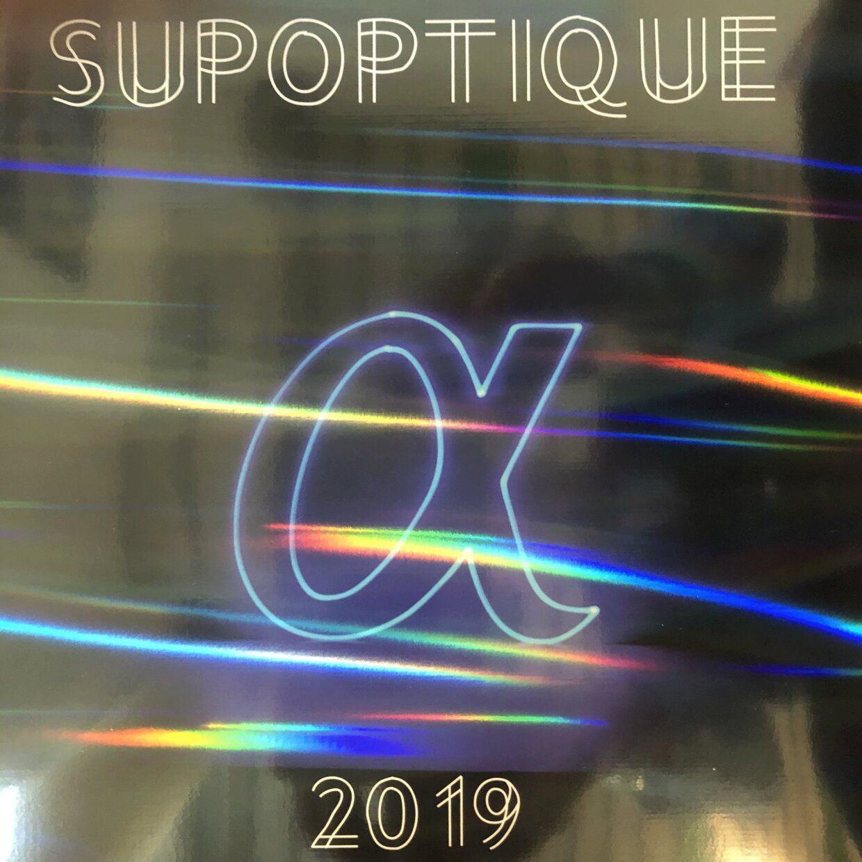 Le texte SUPOPTIQUE au-dessus d'un symbole alpha néon, 2019 en dessous, le tout sur un fond holographique scintillant.
