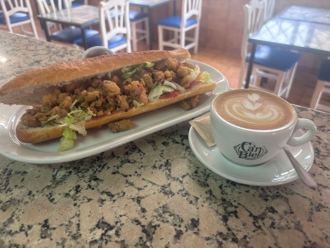 Un sándwich en un plato junto a un café con leche en un platillo en la mesa de un café.