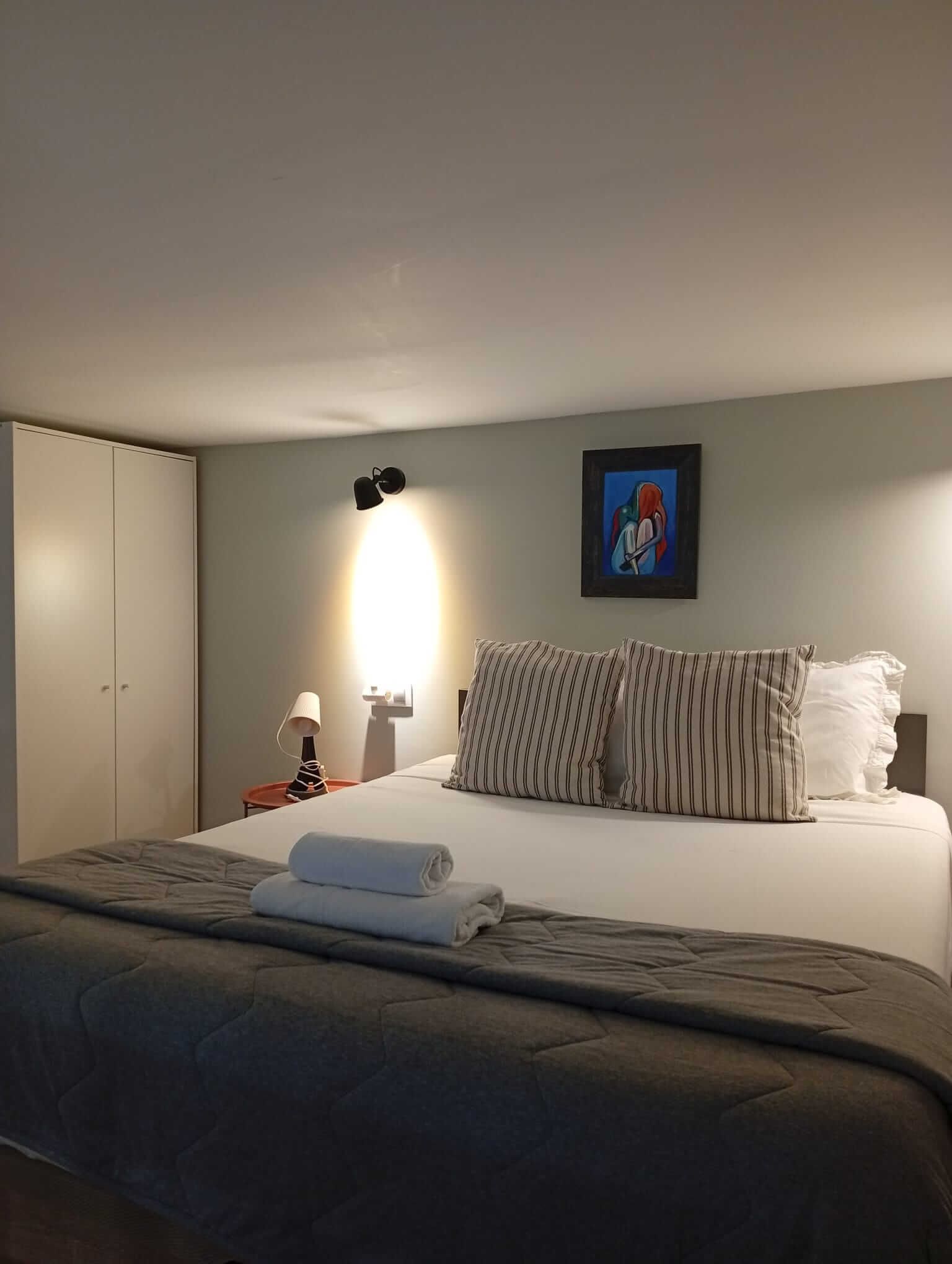 Dormitorio con una cama bien hecha, toallas dobladas, lámparas de noche y un cuadro enmarcado en la pared.