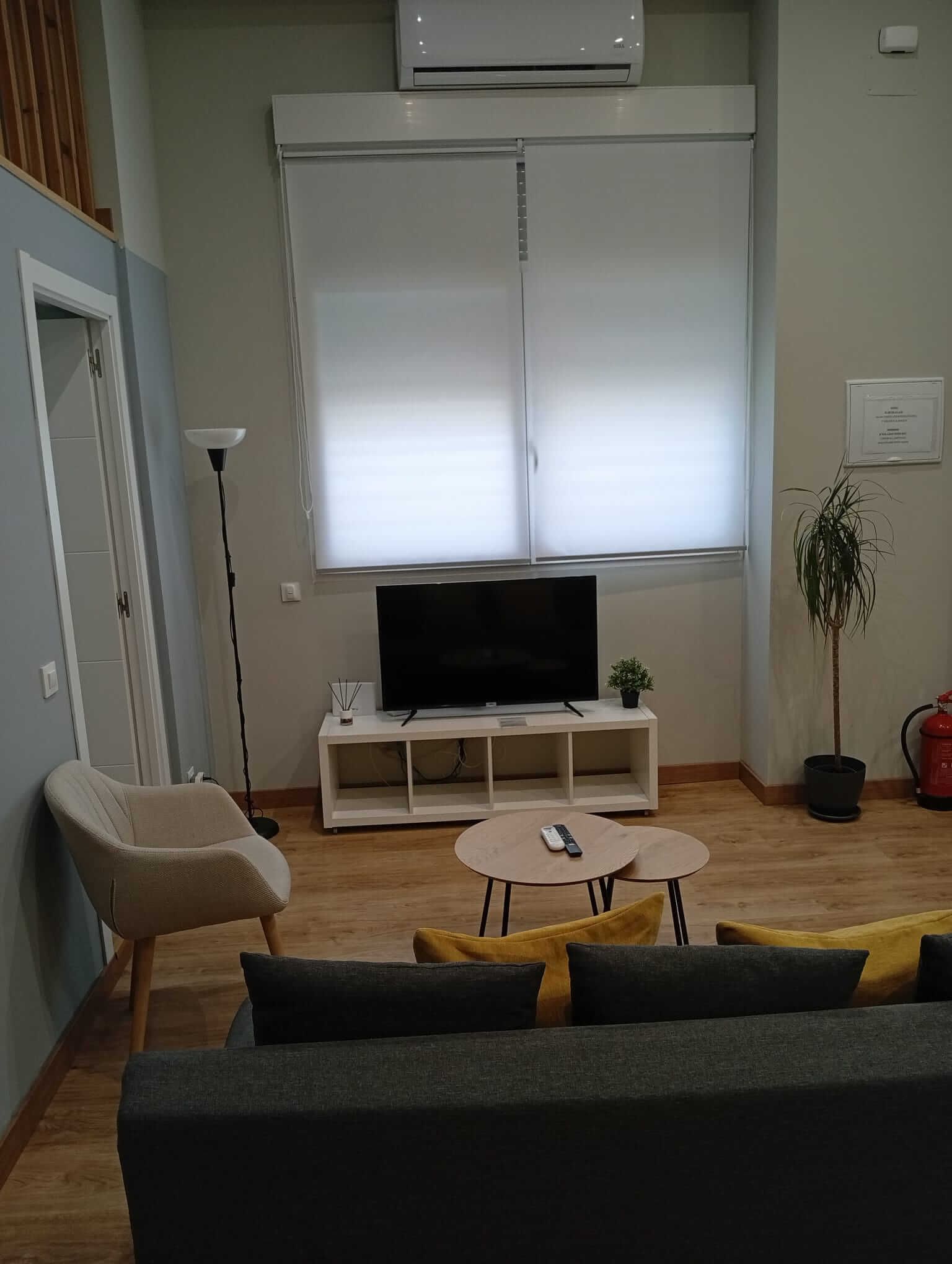 Acogedora sala de estar con sofá, televisor, mesa redonda, planta en maceta y persianas luminosas.