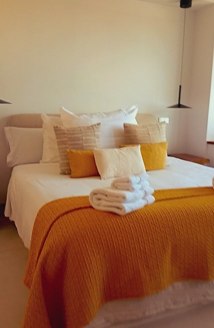 Cama acogedora con almohadas blancas, manta color mostaza y toallas dobladas en una habitación con iluminación tenue.