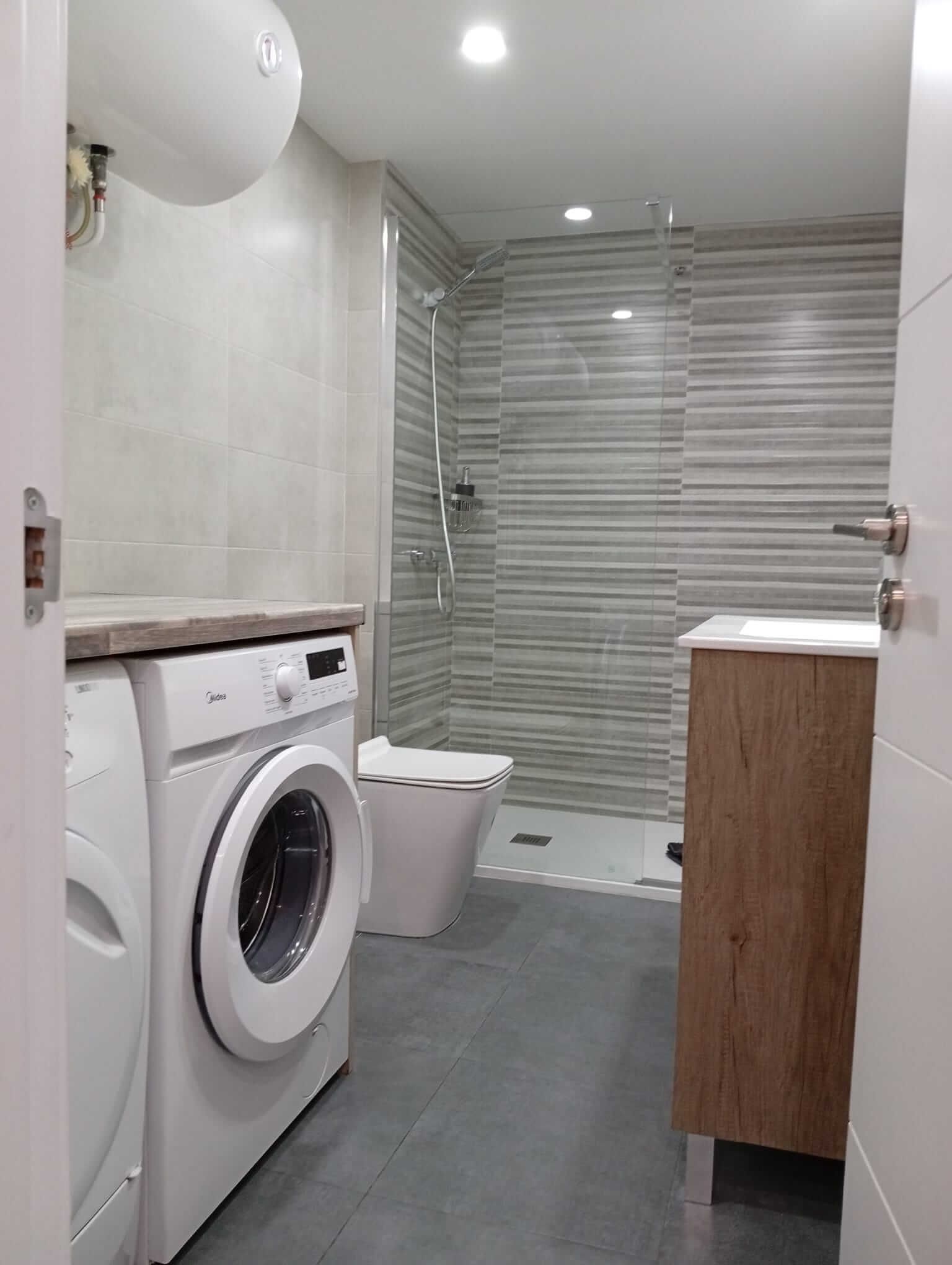 Baño pequeño y moderno con lavadora, inodoro, ducha y tocador de madera en gris y blanco.
