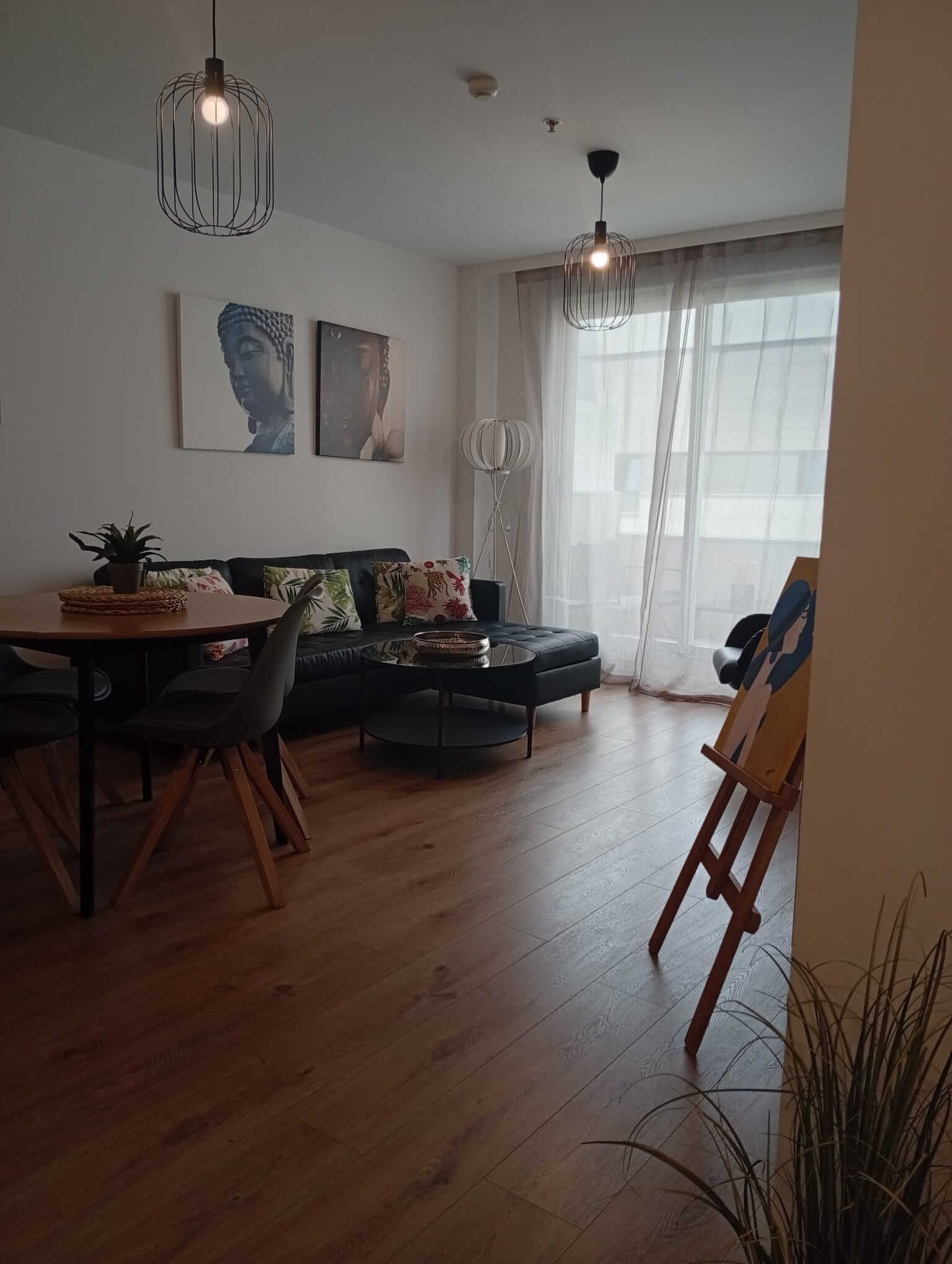 Acogedor salón-comedor con sofá, mesa redonda, lámparas colgantes, cuadros y cortinas transparentes junto a la puerta del balcón.