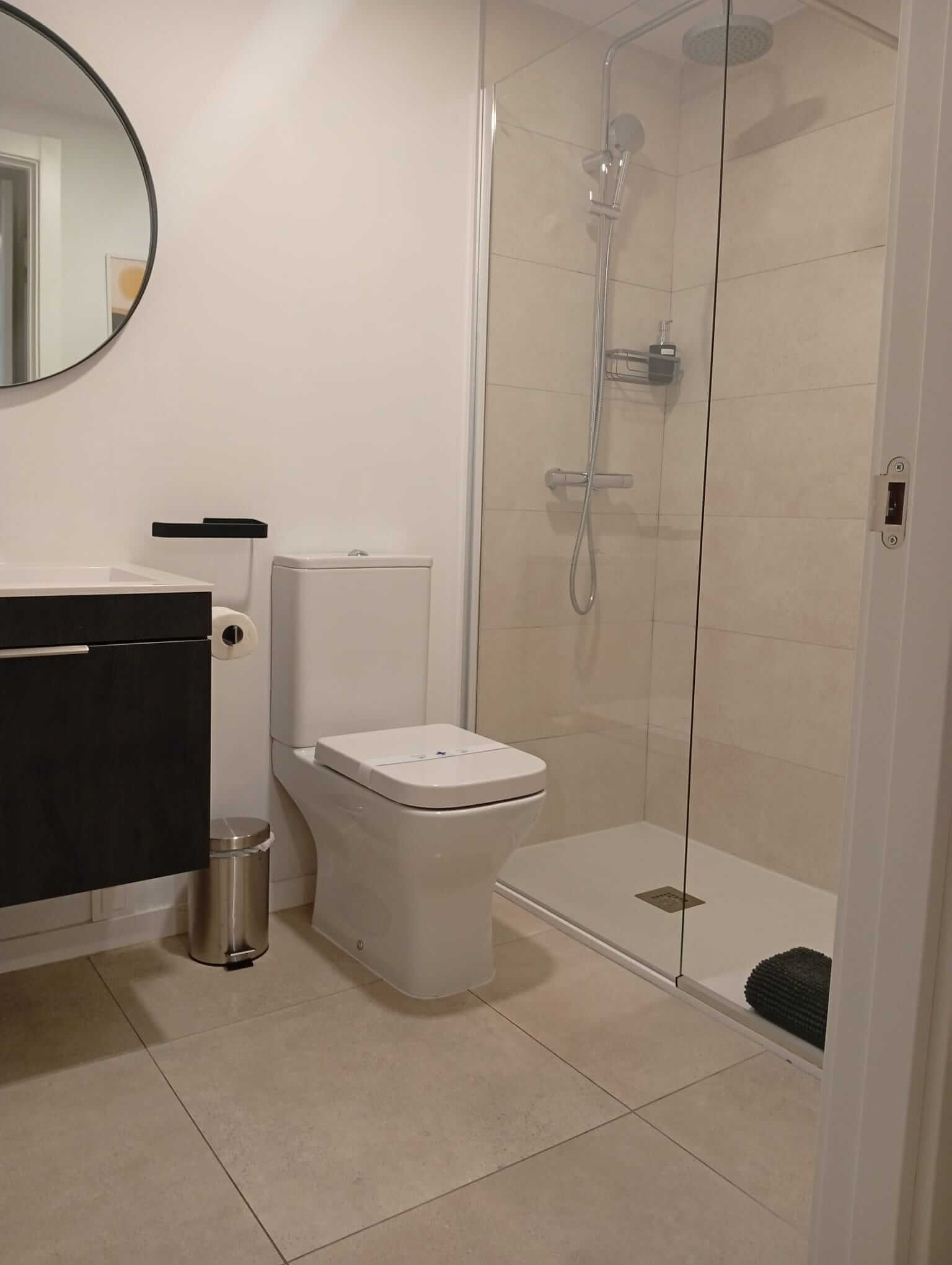 Baño moderno con inodoro, ducha de cristal, suelo de baldosas beige y espejo redondo.