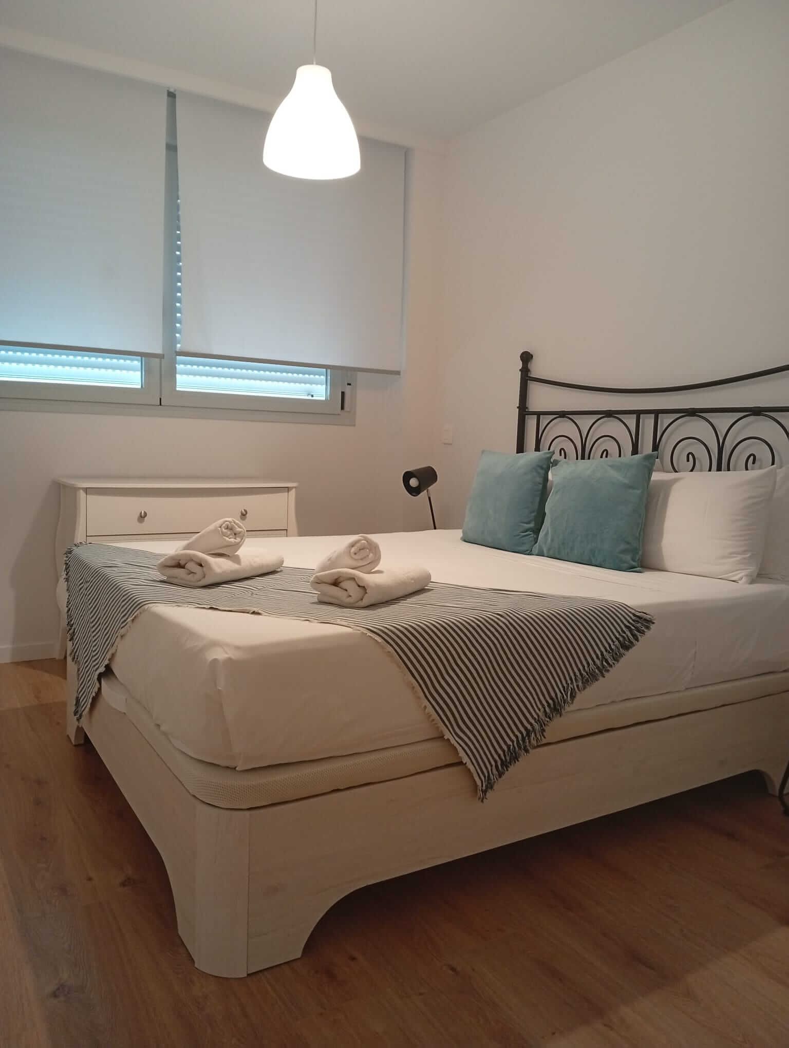 Habitación bien arreglada con cama blanca, almohadas color turquesa, persianas y suelo de madera.