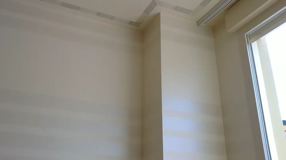 Esquina interior de una habitación con paredes de color blanco roto, molduras y una ventana con persianas.