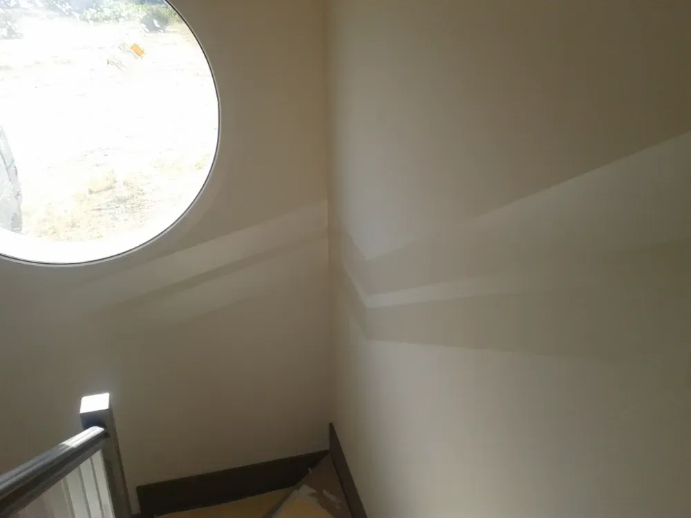 Rincón interior, paredes beige, ventana circular, barandilla, luz solar.