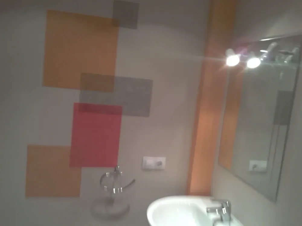 Baño con pared decorada con cuadrados superpuestos de colores, lavabo, espejo y lámpara.