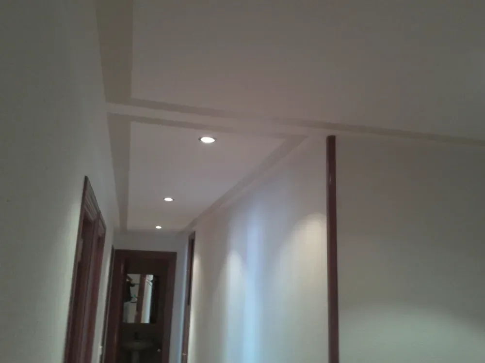 Pasillo con luces empotradas en el techo; paredes blancas, marcos de puertas marrones y una puerta visible.