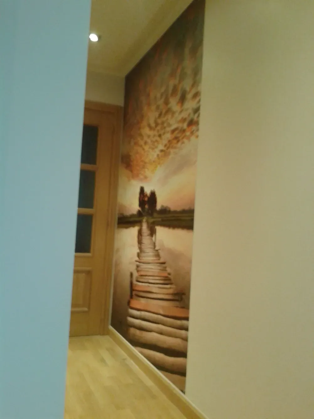 Pasillo estrecho con un mural que representa un puente que se extiende hacia una puesta de sol. Hay una puerta de madera