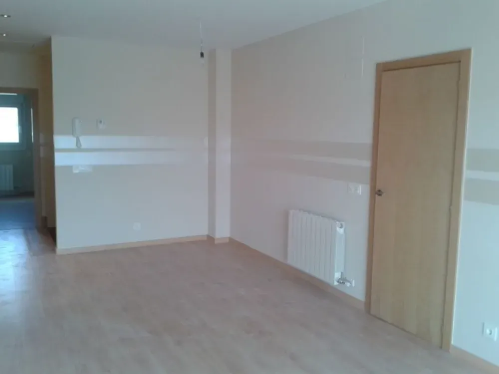 Habitación vacía con suelo de madera clara, paredes beige, una puerta cerrada y un radiador.