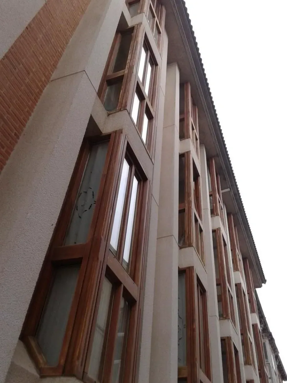 Vista exterior del edificio con altos ventanales con marcos de madera y pilares de hormigón beige.