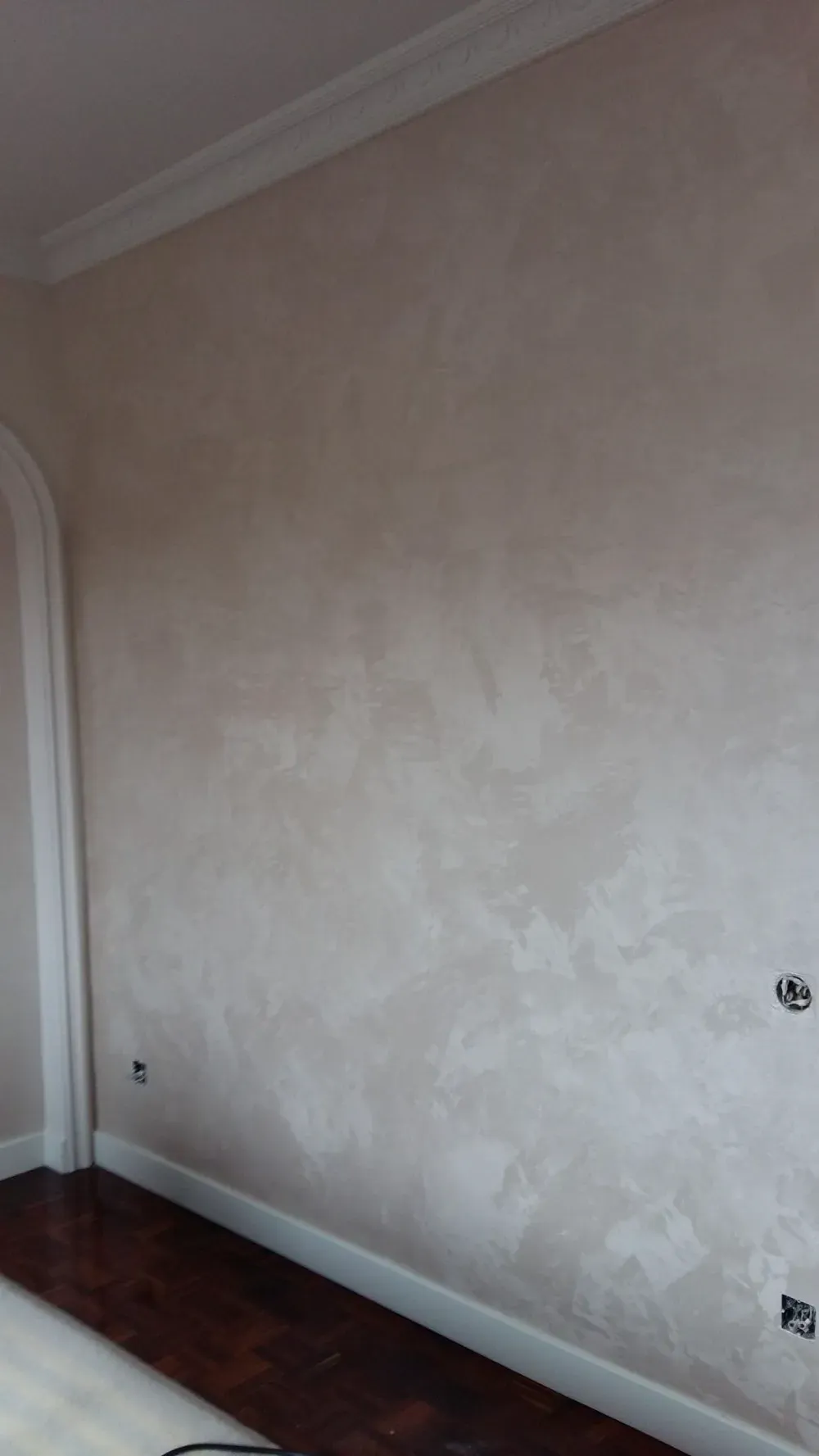 Pared texturizada beige con molduras de corona, piso de madera oscura y enchufes eléctricos.