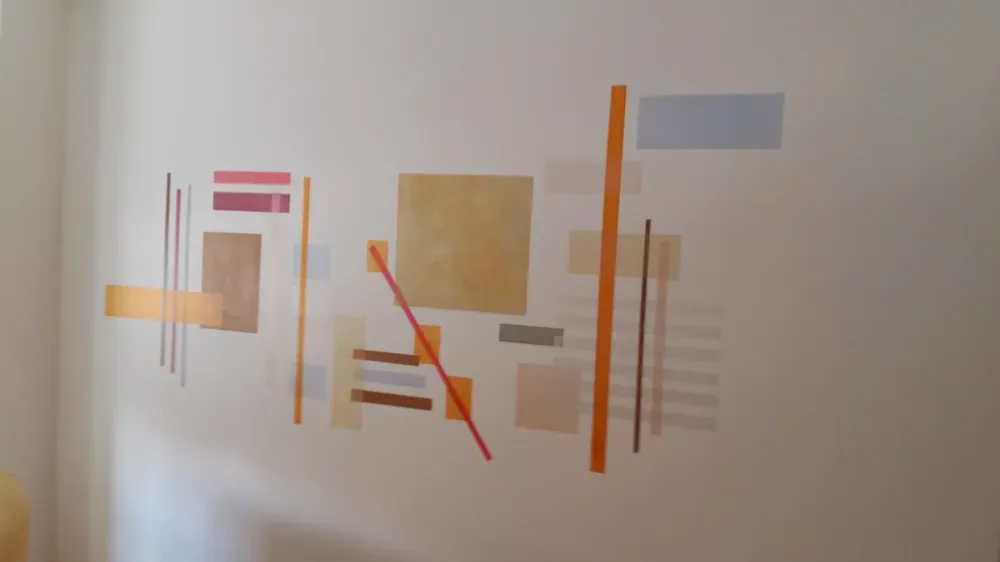 Arte geométrico abstracto sobre una pared blanca, con rectángulos y líneas de color naranja, rojo, marrón y gris.