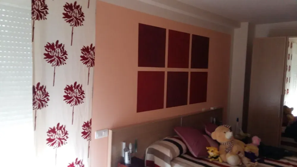 Dormitorio con paredes rosas, cuadros rojos y cortinas. Cama con almohadas, ositos de peluche y edredón de rayas.